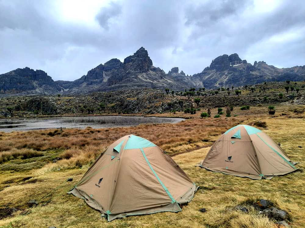 Ahambi Tours camp on the Chogoria Route Mount Kenya — two tents beside a high-altitude tarn with Batian and Nelion peaks rising behind Giant Groundsels on the moorland