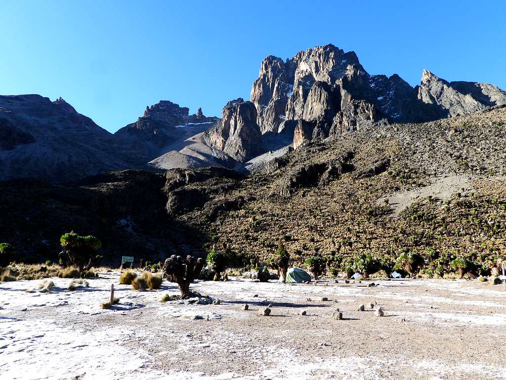 Shipton's Camp at 4,200m on the Sirimon Route Mount Kenya — trekker and tent at the high camp with Batian and Nelion peaks towering above under a deep blue sky