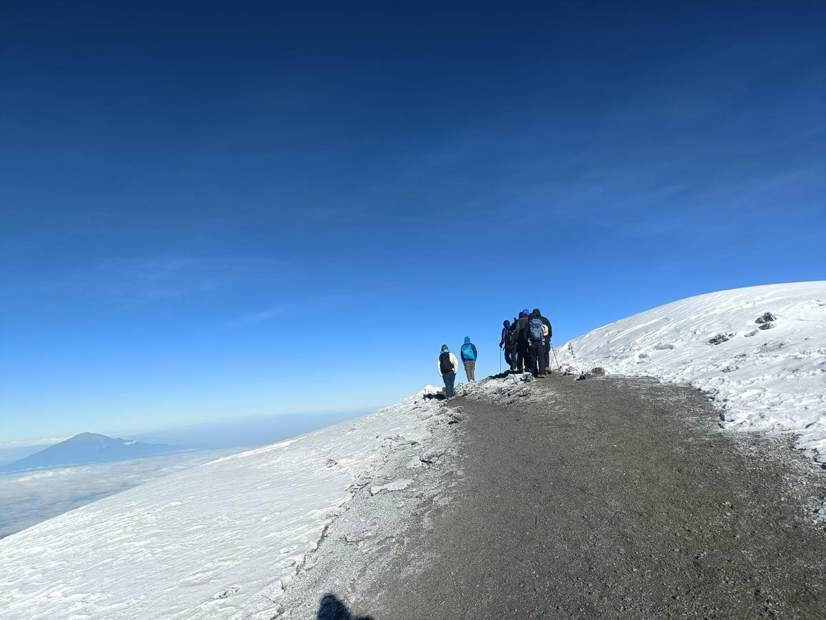 Ahambi Tours trekkers on the Kilimanjaro crater rim approaching Uhuru Peak at 5,895m — above the clouds with glacier and Mount Meru visible